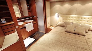 Un dormitorio con una cama grande, cabecero, mesita de noche, espejo, tocador con lavabo y un perchero para toallas.