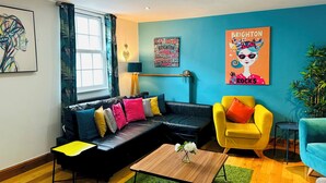 A modern living room with a black leather sofa, a yellow armchair, and a blue armchair. There is a wooden coffee table with a vase of flowers. The room has a blue accent wall and a poster of a woman with the words "Brighton Rocks". - Pebble Mews Free Parking | By My Getaways (Brighton)