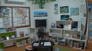A cozy room with a TV, bookshelves, a bulletin board, and a green carpet. - KAGOSHIMA ILCA GUEST HOUSE - Hostel (Kagoshima)