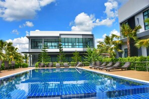 A modern hotel with a swimming pool, lounge chairs, and palm trees.