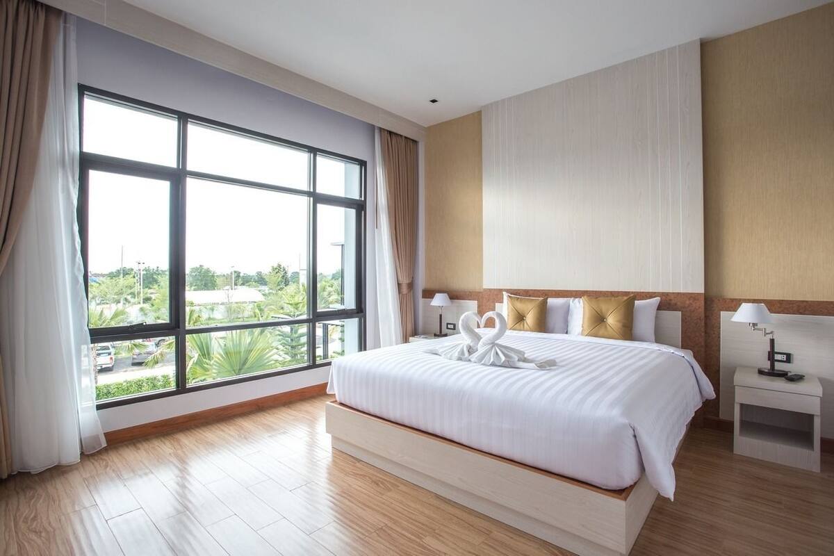 Une chambre d’hôtel avec une grande fenêtre, un lit avec une literie blanche, deux tables de chevet avec des lampes, et une vue sur la verdure à l’extérieur.