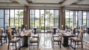 A spacious dining area with tables and chairs, large windows offering views of greenery, and ceiling fans. - Eireann Boutique Hotel (Prakhon Chai)