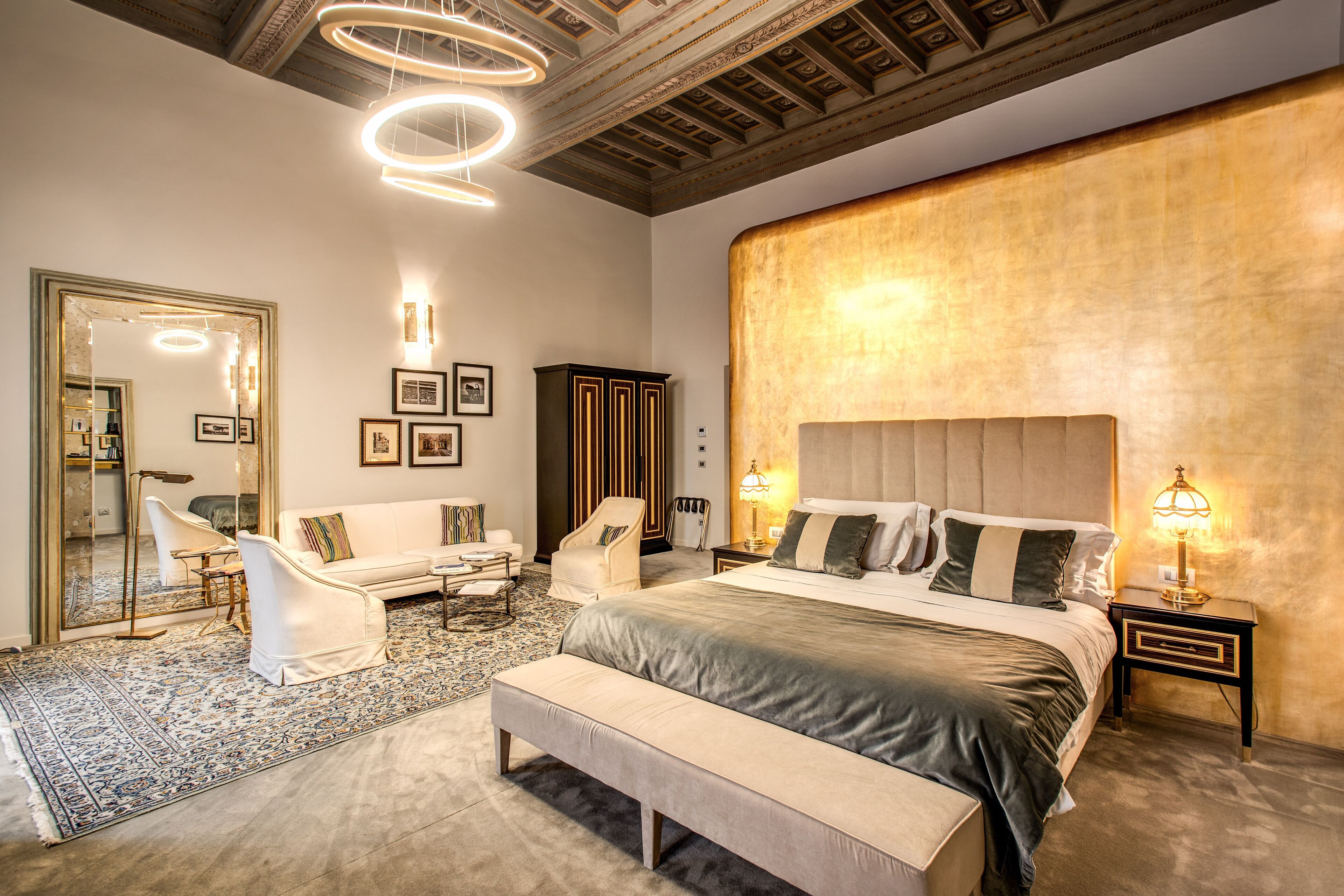 A spacious bedroom with a large bed, a sitting area with a sofa and chairs, a mirror, and a decorative ceiling.