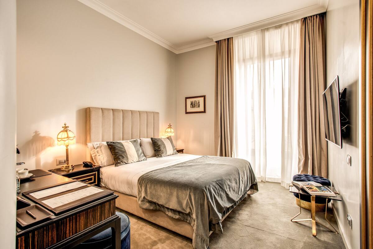 A hotel room with a large bed, bedside lamps, a desk with a lamp, a TV mounted on the wall, and a window with curtains.