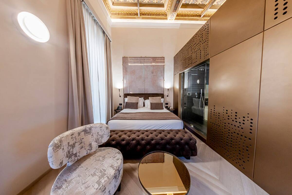 A modern hotel room with a large bed, a seating area with a chair, a mirror, and a wardrobe.