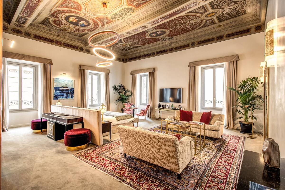 A spacious living room with a large sofa, a coffee table, and a TV. There is a ceiling with intricate designs and a chandelier.