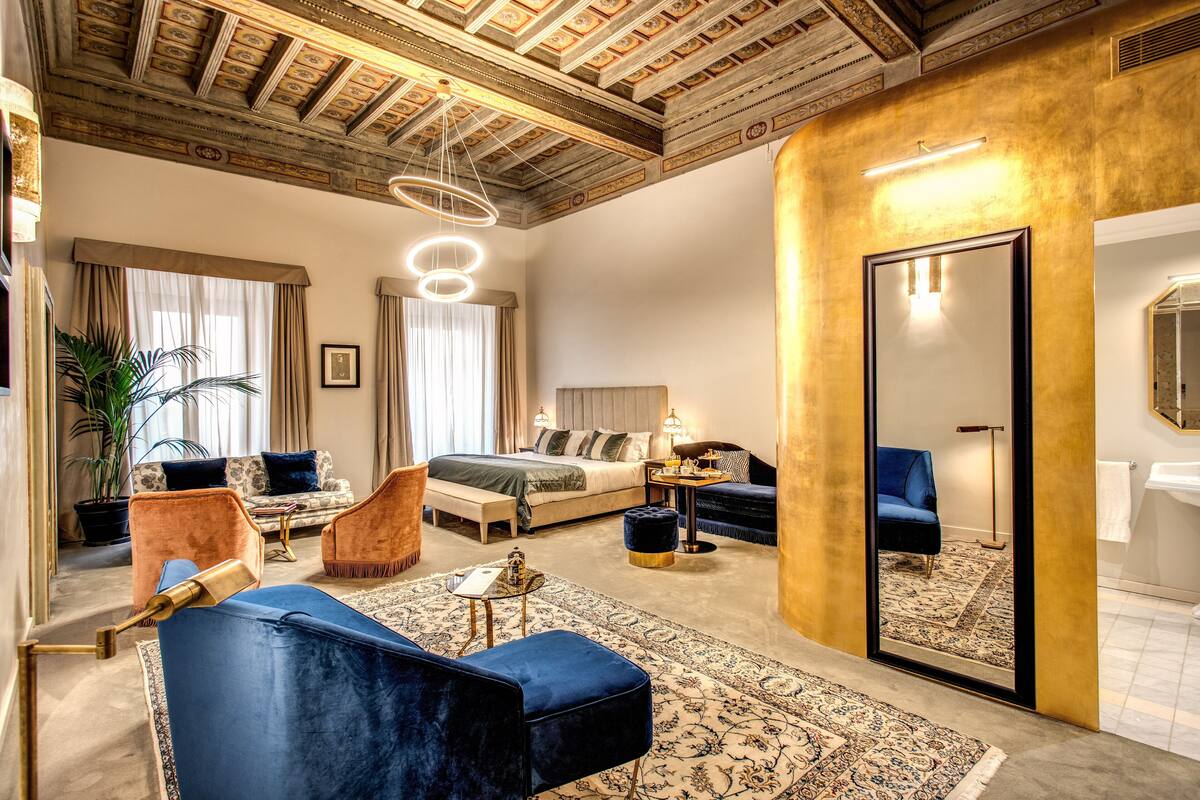 A spacious hotel room with a large bed, a sitting area with a sofa and armchairs, a small table, and a large mirror.