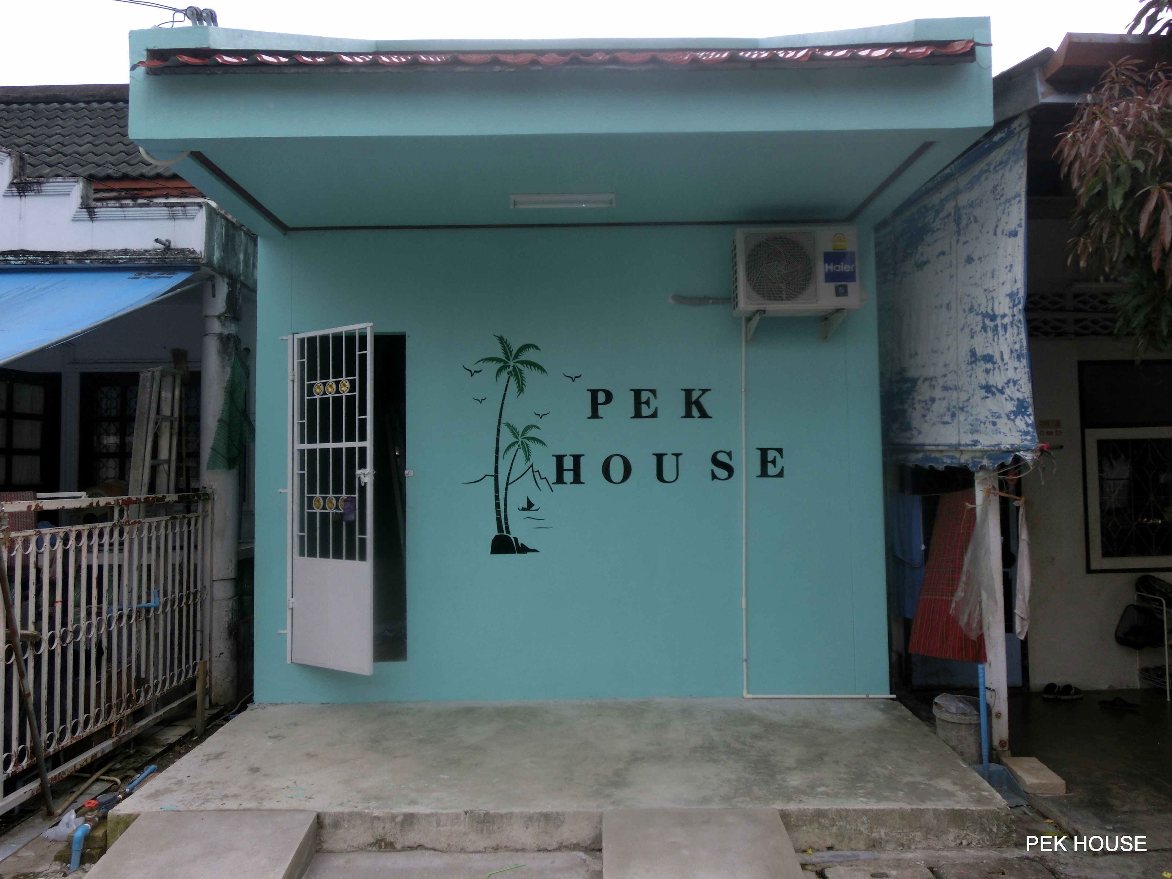 Ein Gebäude mit einer türkisblauen Fassade, einer Tür und einer Wand mit der Aufschrift 'PEK HOUSE' und einer Palmen-Grafik.