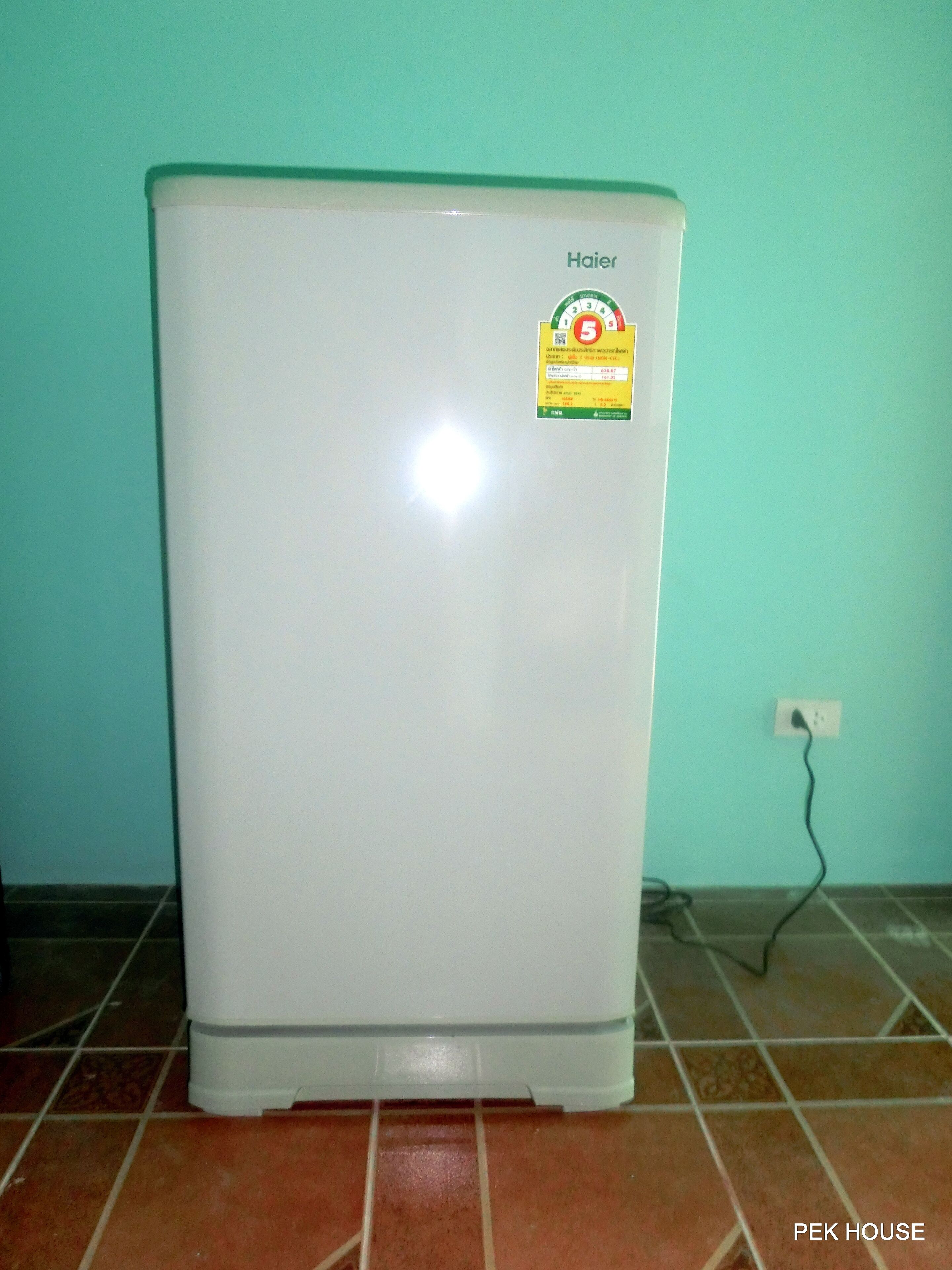 A white refrigerator with a sticker on the front, placed against a teal wall with a tiled floor.