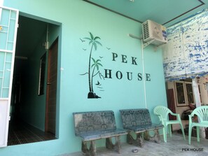 A teal-colored exterior wall with the text "PEK HOUSE" and a palm tree mural. There are two wooden benches and a green plastic chair outside. - Pek House (Phuket)