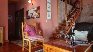 A cozy living room with a wooden chair, a wooden table with wine glasses, a staircase, and a wall with framed pictures. - Artesano III (Navarredonda de Gredos)