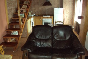 A living room with a black leather sofa, wooden staircase, kitchen area, and brick walls.