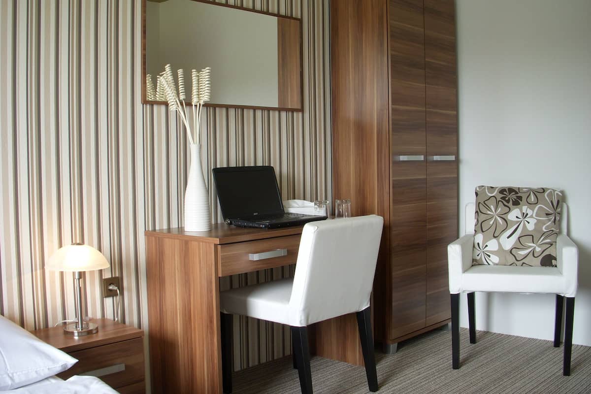 A hotel room with a wooden desk, a laptop, a white chair, a bedside table with a lamp, and a wardrobe.