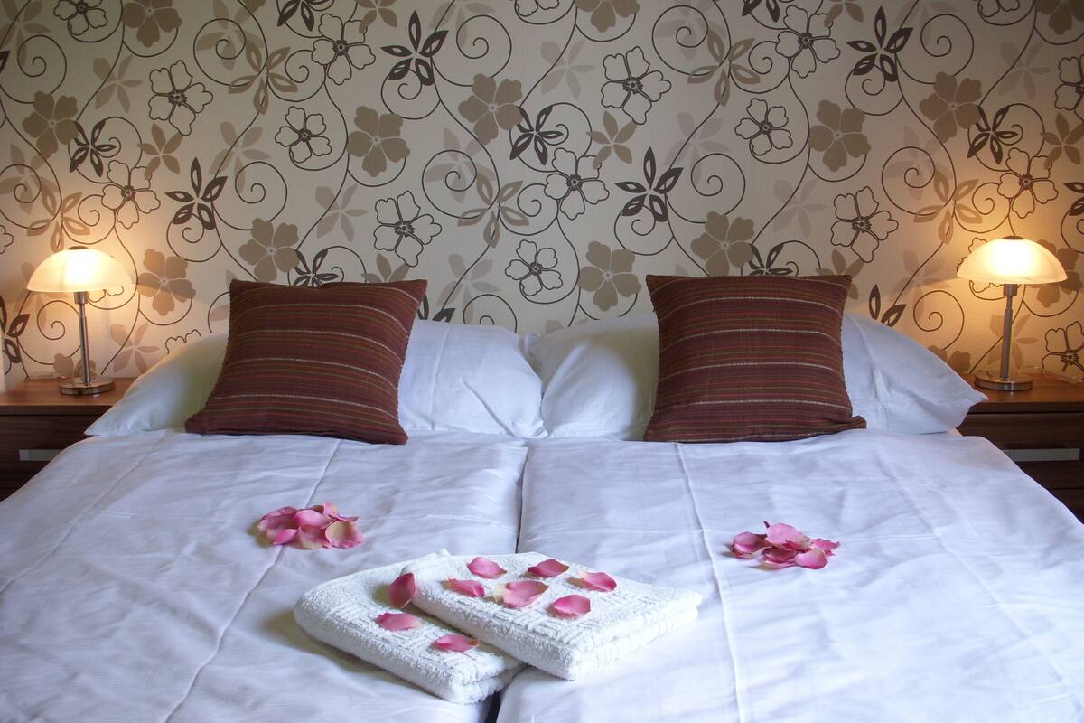 A neatly made bed with white linens, two pillows, and a pair of towels with rose petals.