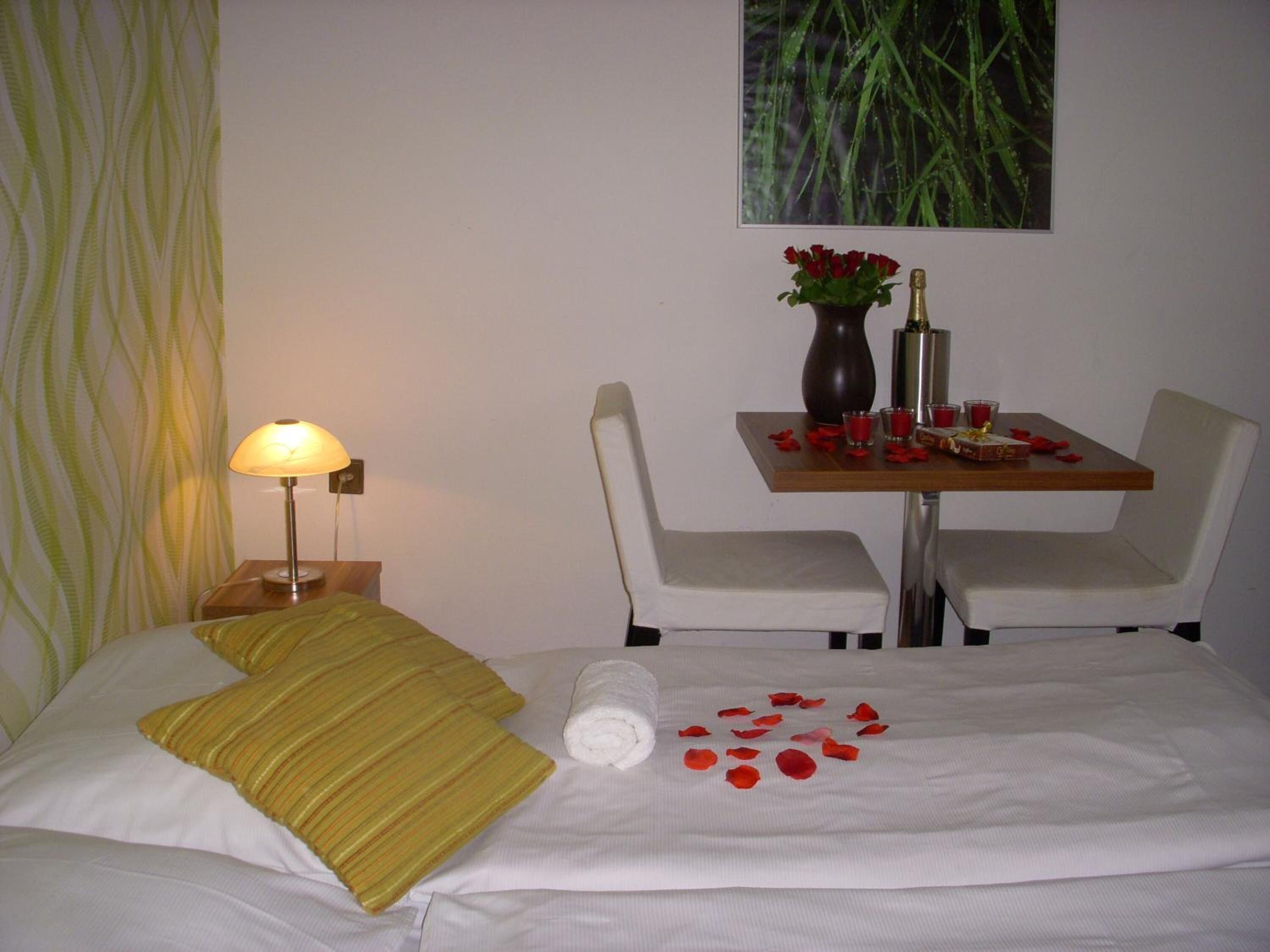Habitación de hotel con cama, mesita de noche con lámpara, una pequeña mesa con un jarrón de flores y una silla.