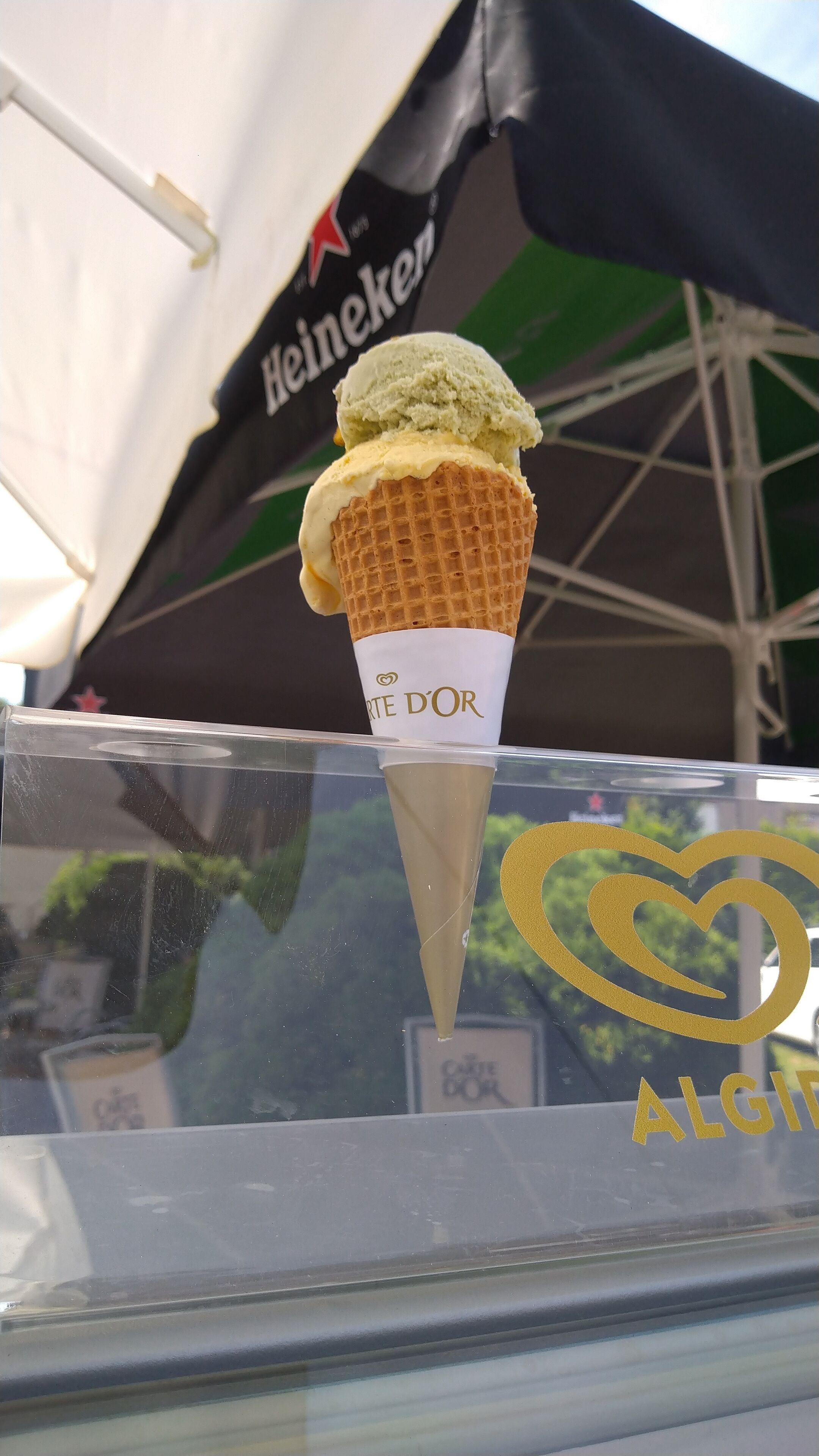 Un cono de helado con un cono de gofre y una taza de papel blanca con la etiqueta "ART D'OR" frente a una ventana de vidrio.
