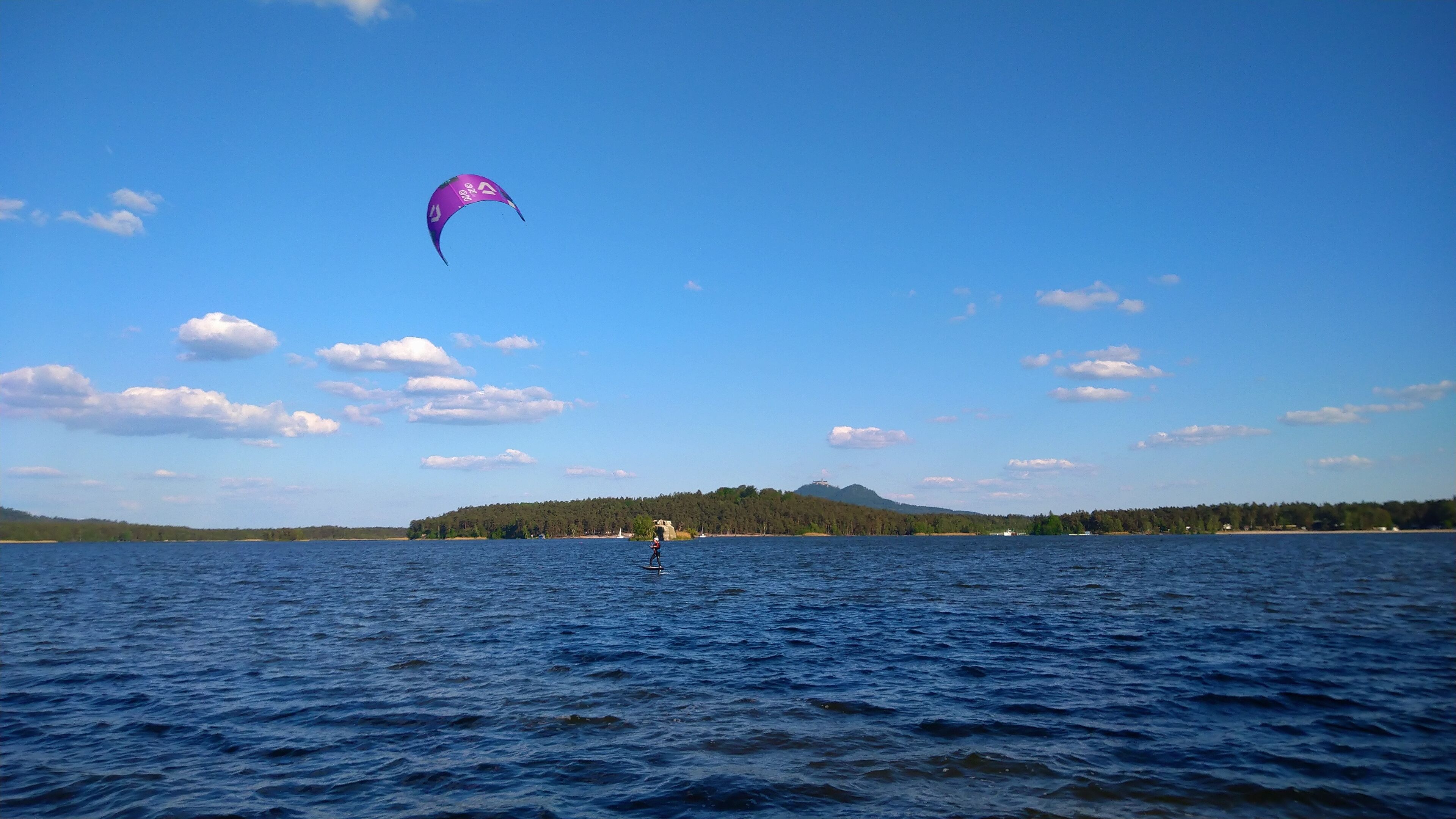 A person kiteboarding on a lake with a clear sky and distant trees.