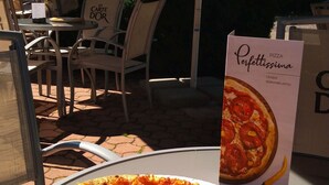 A pizza on a table with a sign that reads "Perfettissima" in an outdoor seating area with chairs and umbrellas.