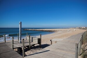 A wooden boardwalk leading to a sandy beach with a rocky breakwater in the background.