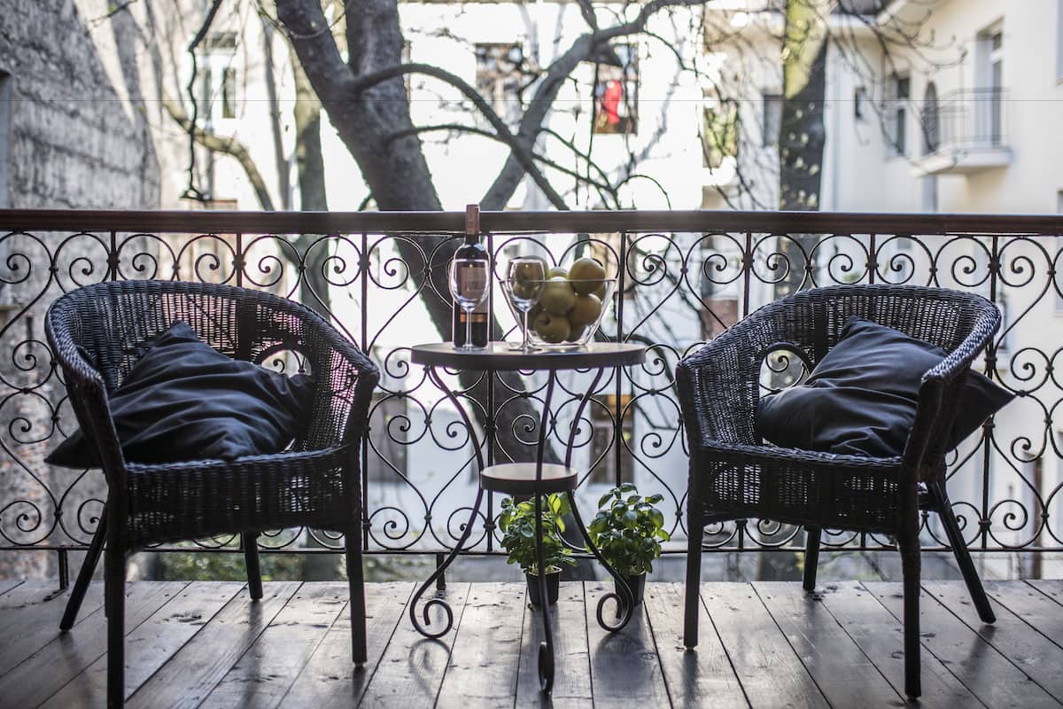 Two wicker chairs with cushions on a wooden deck, a small table with a bottle and a bowl of fruit, and potted plants.
