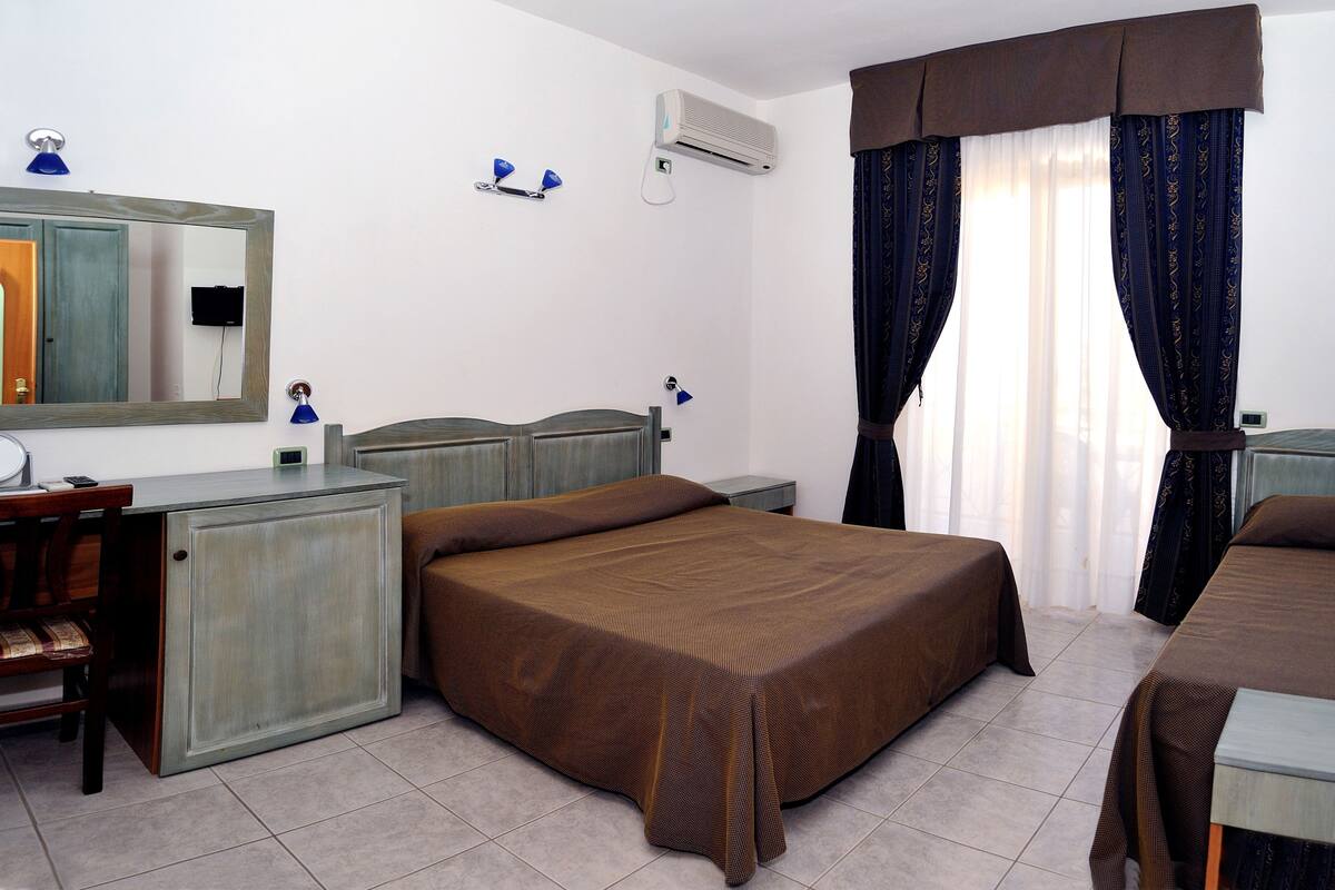 Habitación de hotel con cama, escritorio, silla, espejo, aire acondicionado y ventana con cortinas.