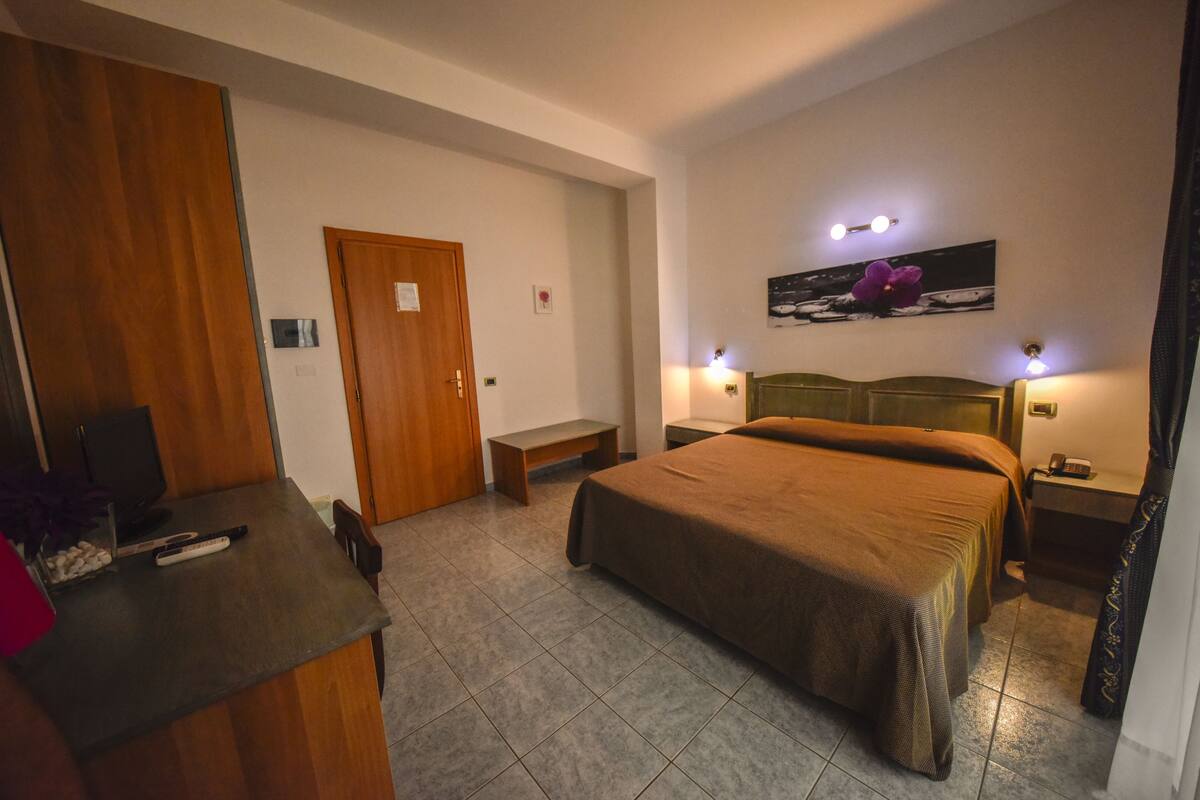 Habitación de hotel con una cama grande, un escritorio con computadora, un banco y una puerta.
