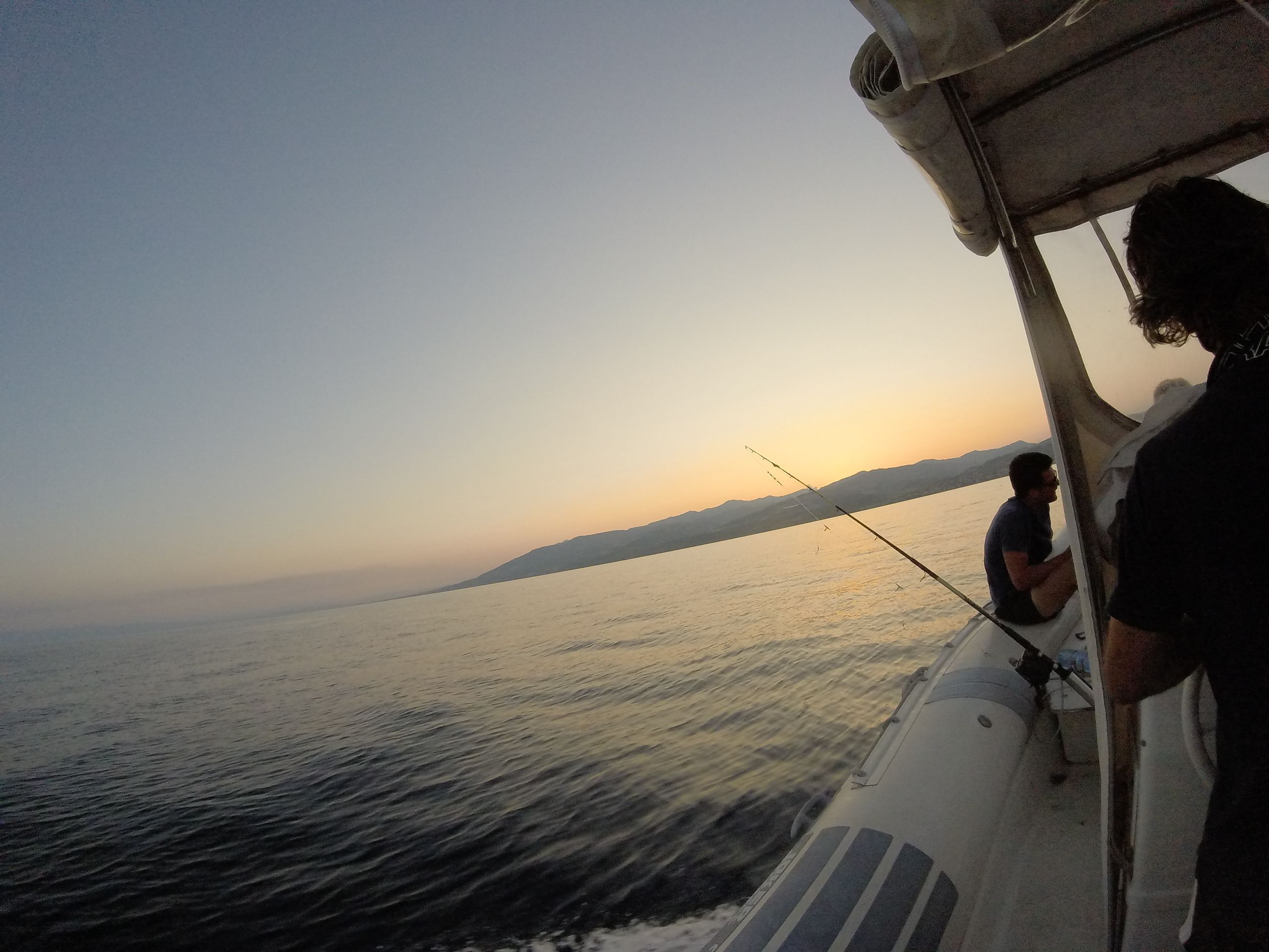 Un bote con dos personas pescando, agua tranquila y una orilla distante al atardecer.