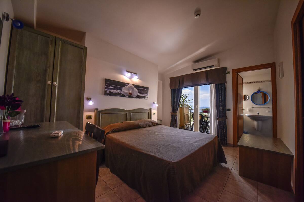 Habitación de hotel con cama, escritorio, silla, armario y baño con lavamanos y espejo.