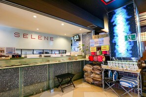 A hotel reception area with a 'SELENE' sign, a glass counter, a small stool, and various amenities like a TV, plants, and a display of bottled water. - Hotel Chuo Selene (Osaka)