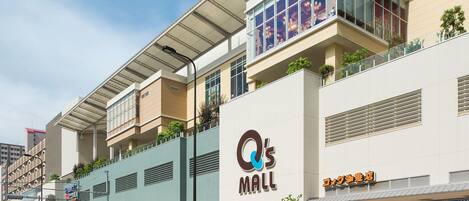 Un centro comercial moderno con un llamativo letrero que dice 'Q's Mall', asientos al aire libre y diversas tiendas.