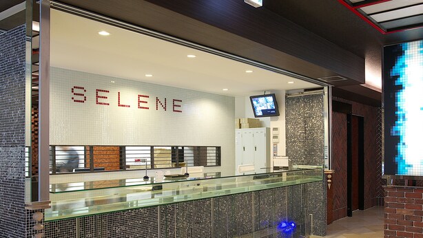 A reception area with a glass counter, a tiled wall with the name 'SELENE', a television screen, and a security camera. - Hotel Chuo Selene (Osaka)