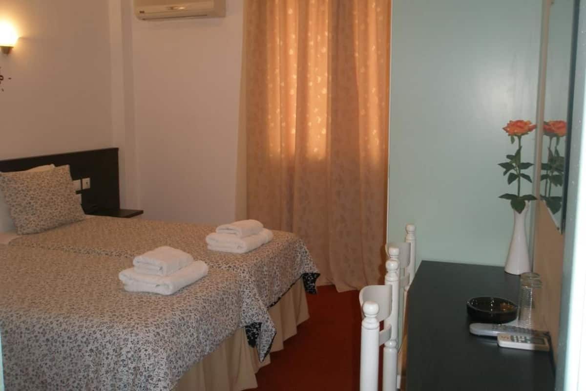 A hotel room with a bed, a bedside table with a remote control, a vase with a rose, and a window with curtains.