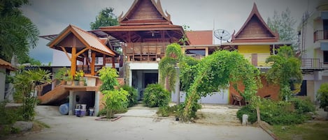 Una casa de madera tradicional con una estructura central elevada, rodeada de abundante vegetación y un patio empedrado.