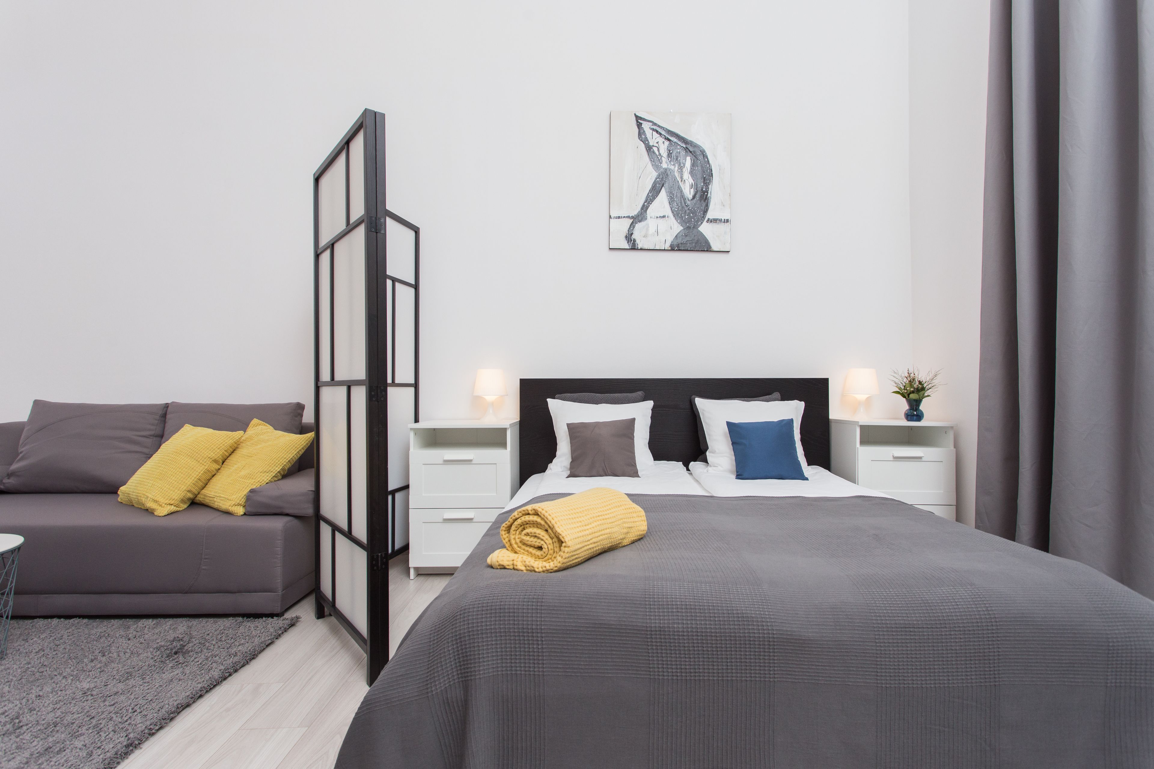 A modern bedroom with a grey sofa, a large bed with a rolled-up blanket, two bedside tables with lamps, and a wall-mounted artwork.