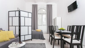 A modern living room with a sofa, a dining table with chairs, a TV mounted on the wall, and a large window with curtains.