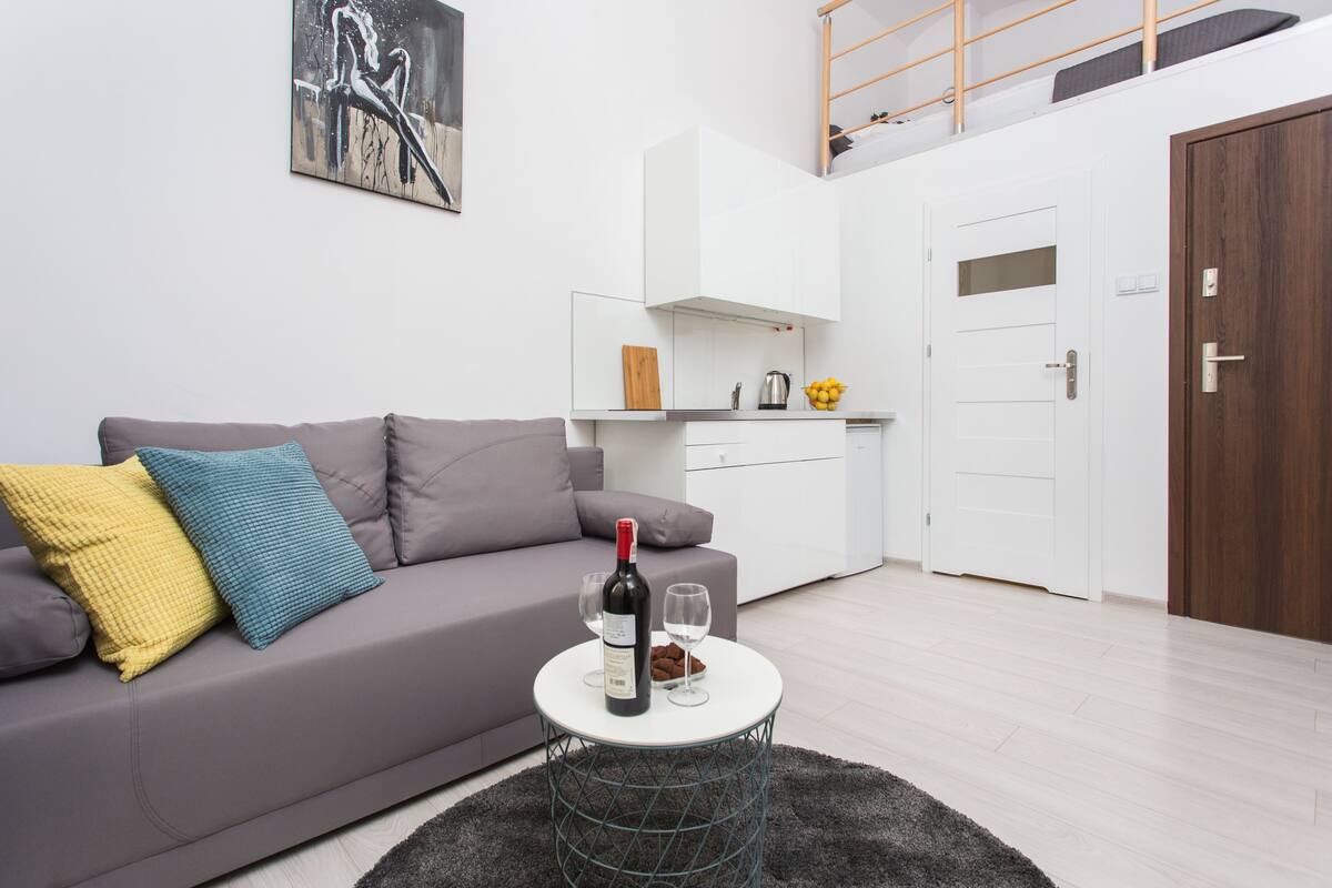 Uma sala de estar moderna com um sofá cinzento, uma pequena mesa redonda com uma garrafa e copos, uma área de cozinha com armários brancos e uma porta de madeira.