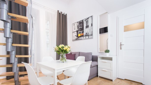 A modern living room with a white sofa, a dining table with chairs, a staircase, and a painting on the wall.