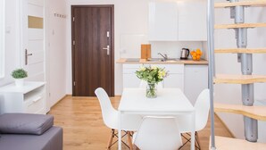 A compact living space with a loft bedroom, a dining area with a white table and chairs, a kitchen with white cabinets, and a wooden staircase.