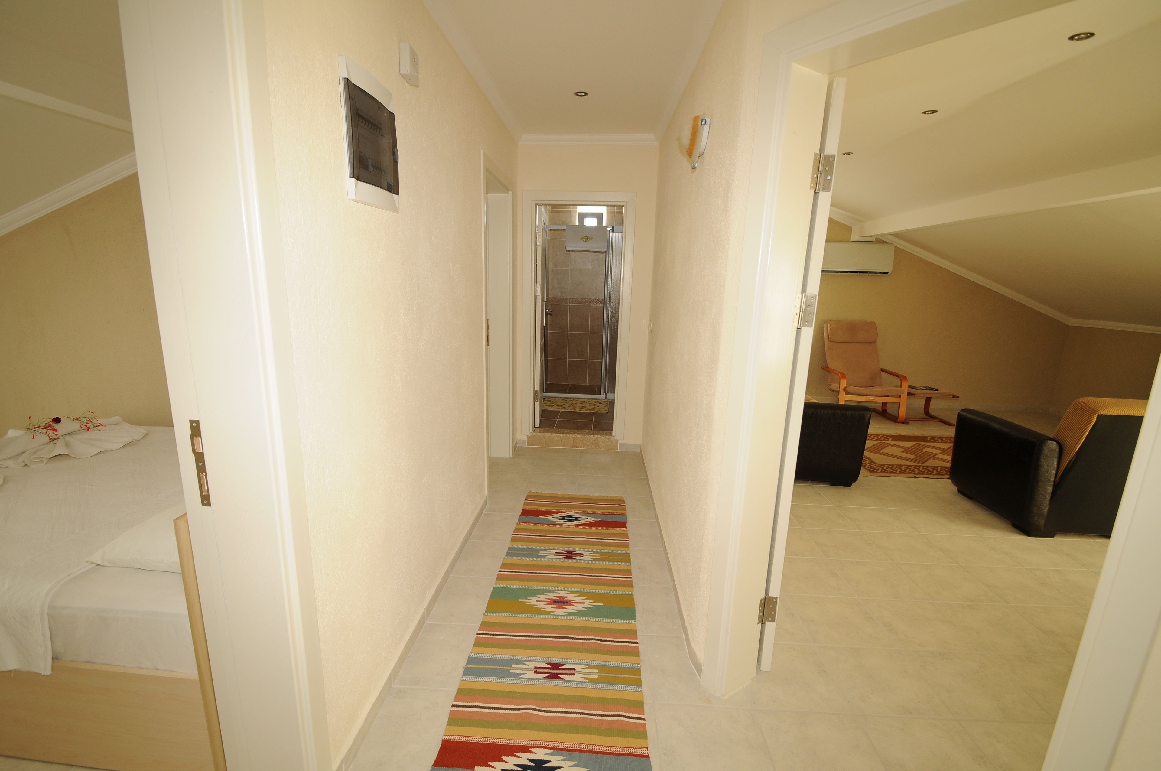 A narrow hallway with a colorful rug, a bed on the left, and a seating area with a chair and ottoman on the right.