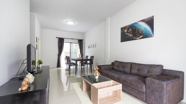A modern living room with a grey sofa, a coffee table, a flat-screen TV, and a dining area with a table and chairs. - Supirum Cozy Home 3 Bedroom near Beach (Thep Krasattri)