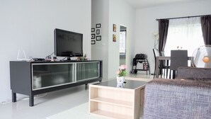A modern living room with a black entertainment unit, a glass coffee table, a grey sofa, and a dining area with a fan and a baby stroller. - Supirum Cozy Home 3 Bedroom near Beach (Thep Krasattri)