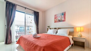A hotel room with a large bed, a nightstand with a lamp, a window with curtains, and a painting on the wall. - Supirum Cozy Home 3 Bedroom near Beach (Thep Krasattri)