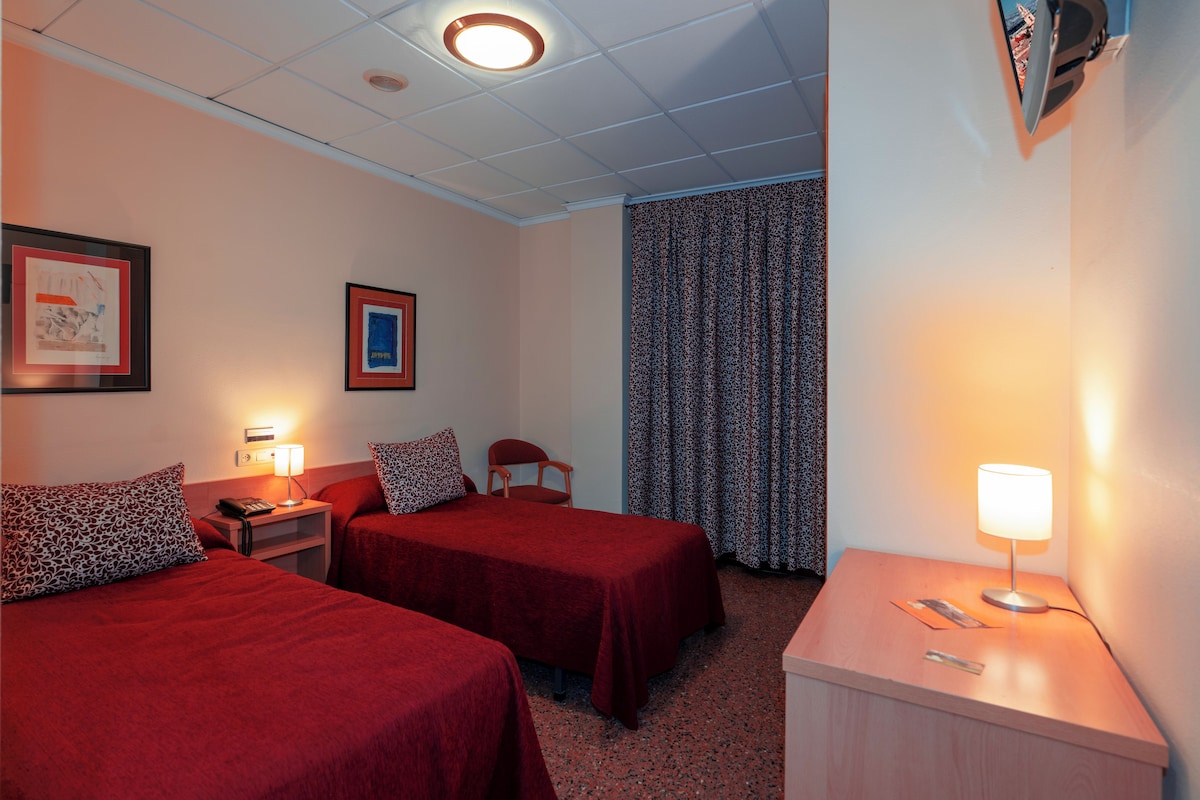 A hotel room with two beds, a desk, a chair, a lamp, and a curtain.