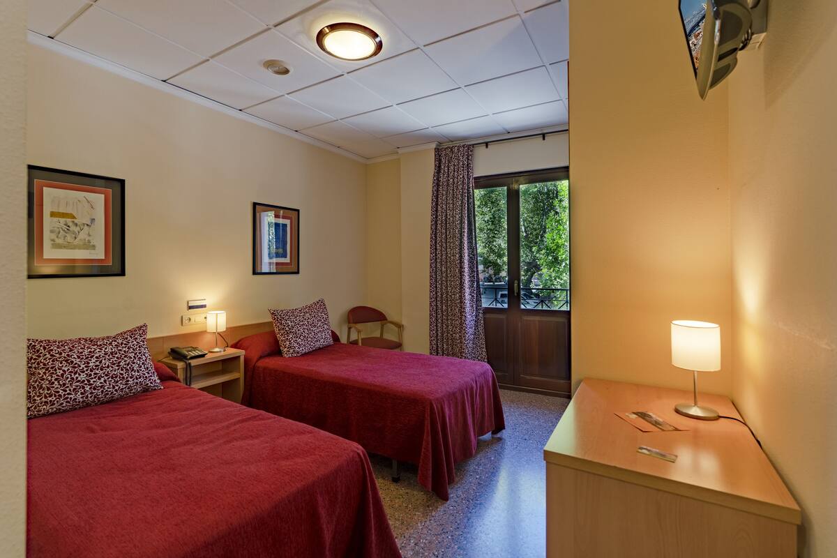 Habitación de hotel con dos camas, una mesita de noche, una lámpara y una ventana con cortinas.