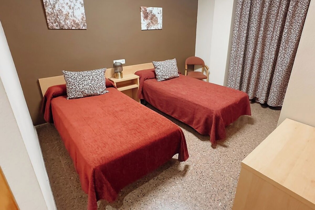 Quarto com duas camas de solteiro, cada uma com colcha vermelha e travesseiro estampado com padrão de leopardo. Uma mesa de cabeceira de madeira com abajur fica entre as camas, e uma cadeira de madeira à direita. A parede é marrom, com duas molduras. Uma cortina estampada é parcialmente visível à direita.