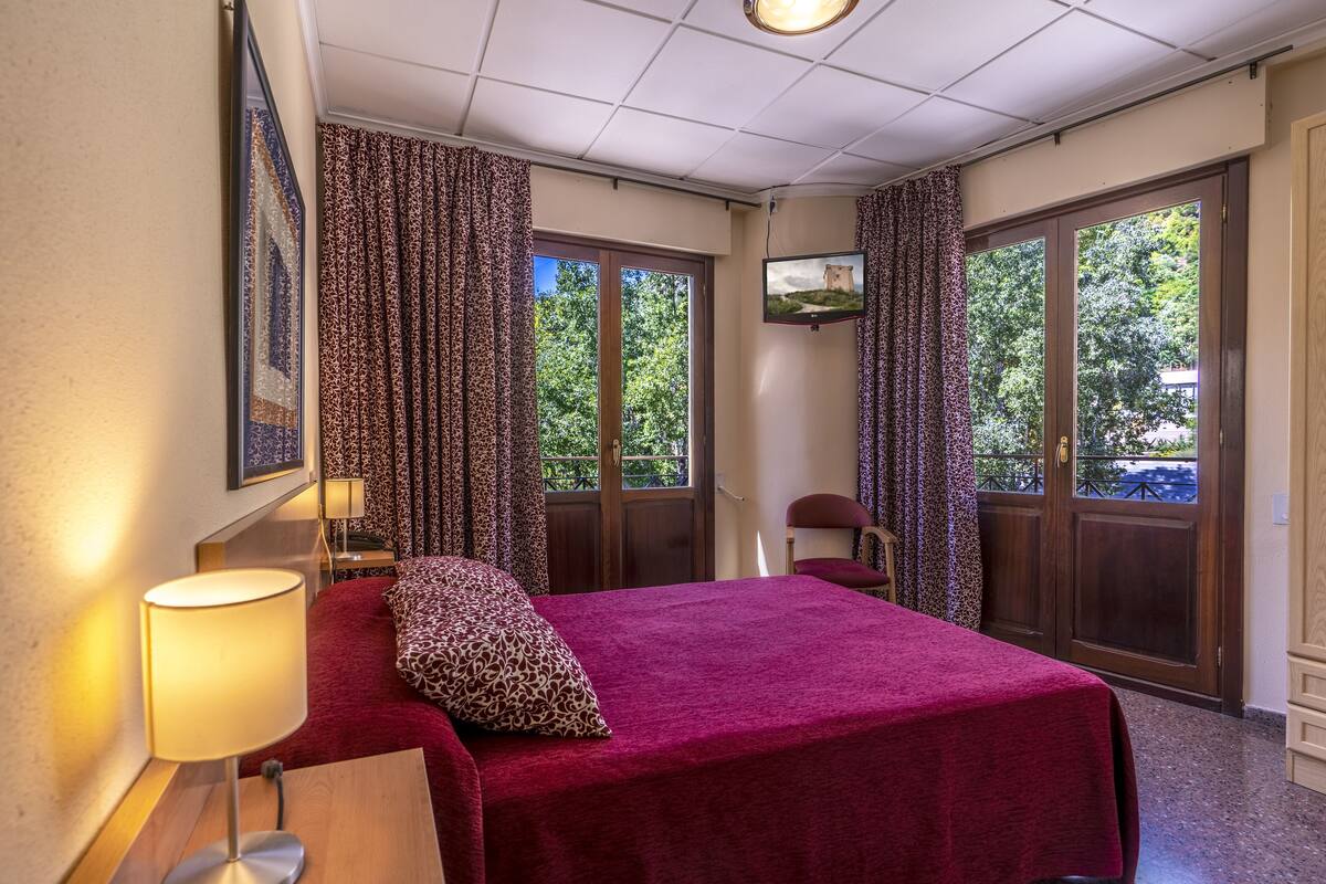 Habitación de hotel con cama, escritorio, silla, televisor y ventanas grandes con cortinas.