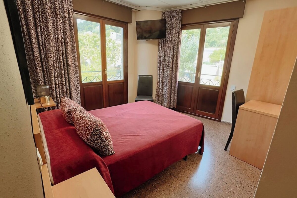 A bedroom with a large bed, a TV mounted on the wall, and a balcony with curtains.