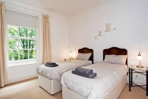 Une chambre avec deux lits, une fenêtre avec des rideaux, une table de chevet avec une lampe et un luminaire fixé au mur.