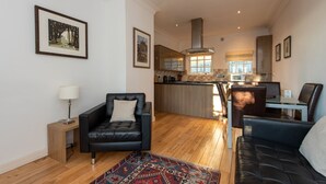 A modern living room with a black leather sofa, a patterned rug, and a wooden coffee table. A kitchen with a large island and a dining area with a glass table and chairs is visible in the background. - Toast Lettings | Apartment 4 The Grove minutes walk from Durham City (Durham)
