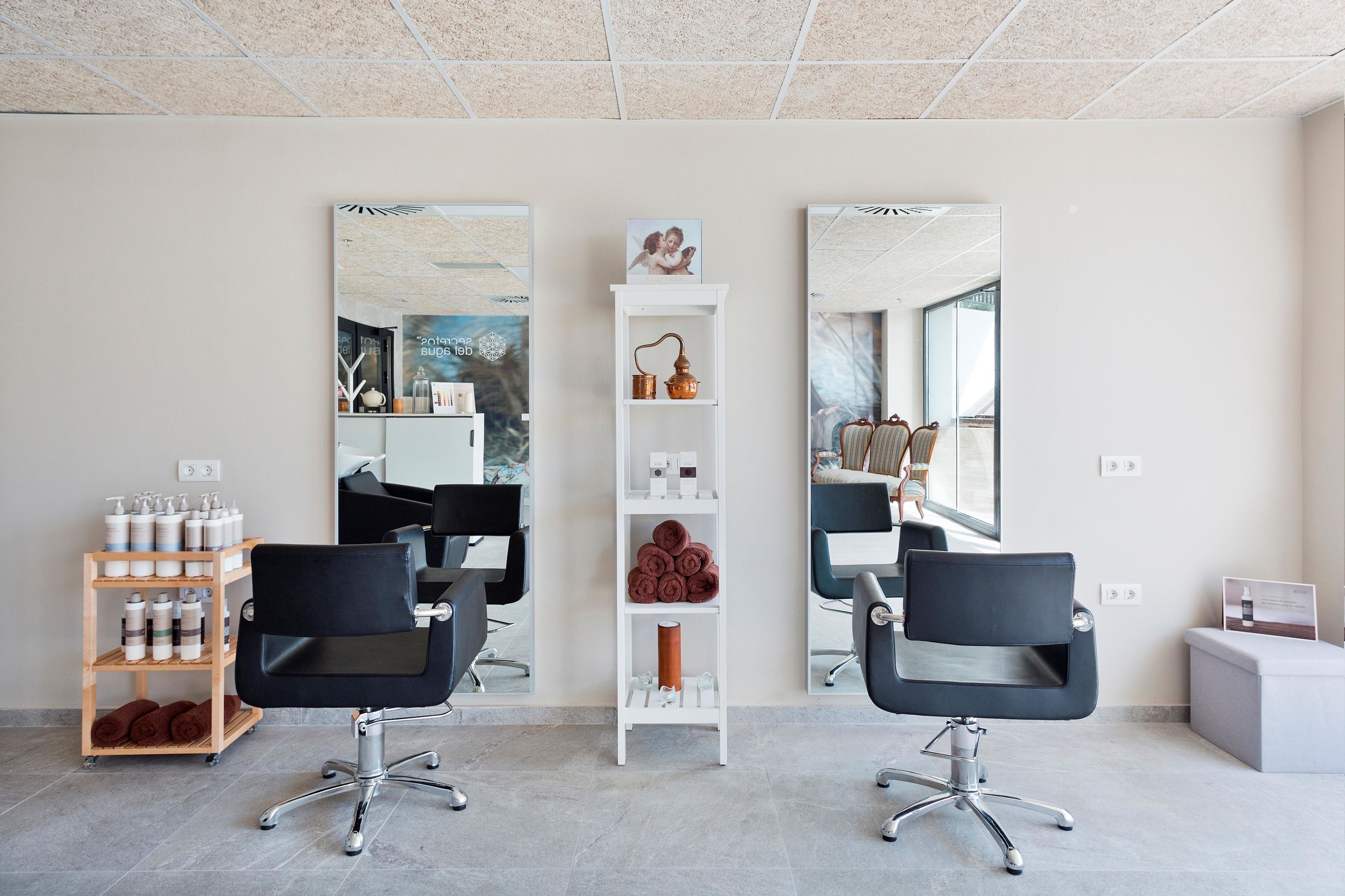 A modern salon with two black salon chairs, a white shelf with hair products, and large mirrors on the walls.
